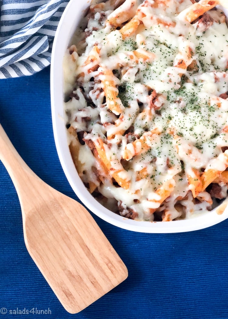 Penne pasta with hearty meat sauce with melted mozzarella cheese in a white casserole dish on a blue background