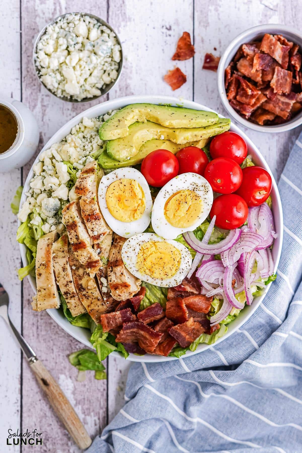 White bowl filled with cobb salad, layered with chicken, bacon, red onion, blue cheese, hard boiled eggs and cherry tomatoes on a blue and white striped napkin.