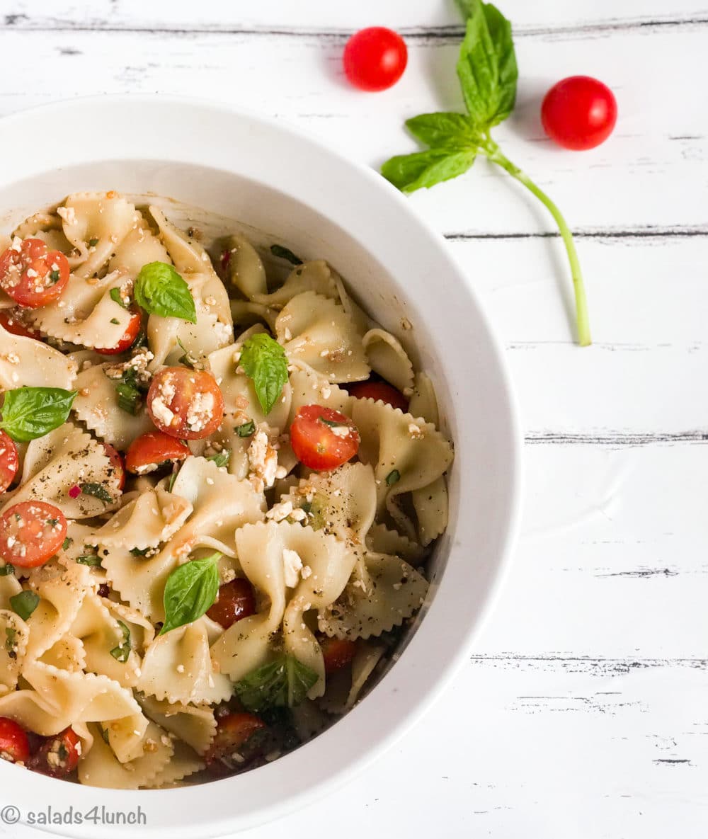 White bowl of bow tie feta bruschetta pasta salad with sprig of basil and roma tomatoes.