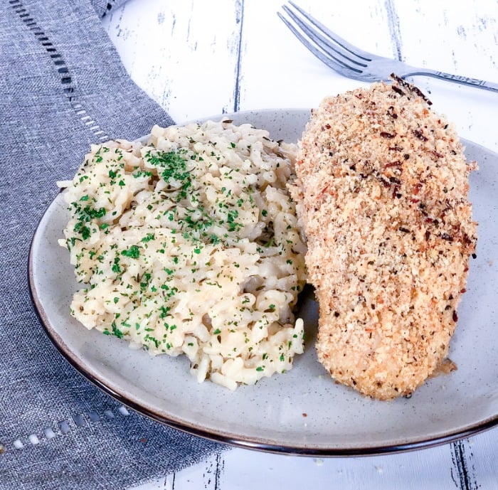 Close up of garlic parmesan baked chicken breast with a side of risotto on a white plate on a grey napkin.