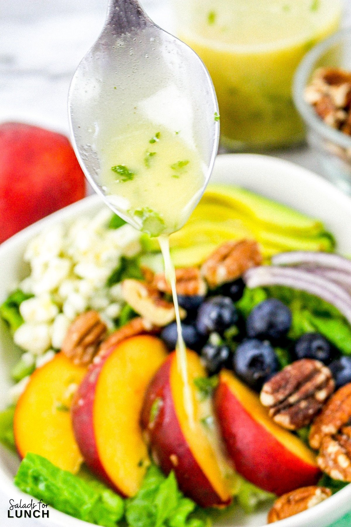 Fresh mint salad dressing being drizzled from a spoon onto a summer peach salad with blueberries, avocado, onions and blue cheese.