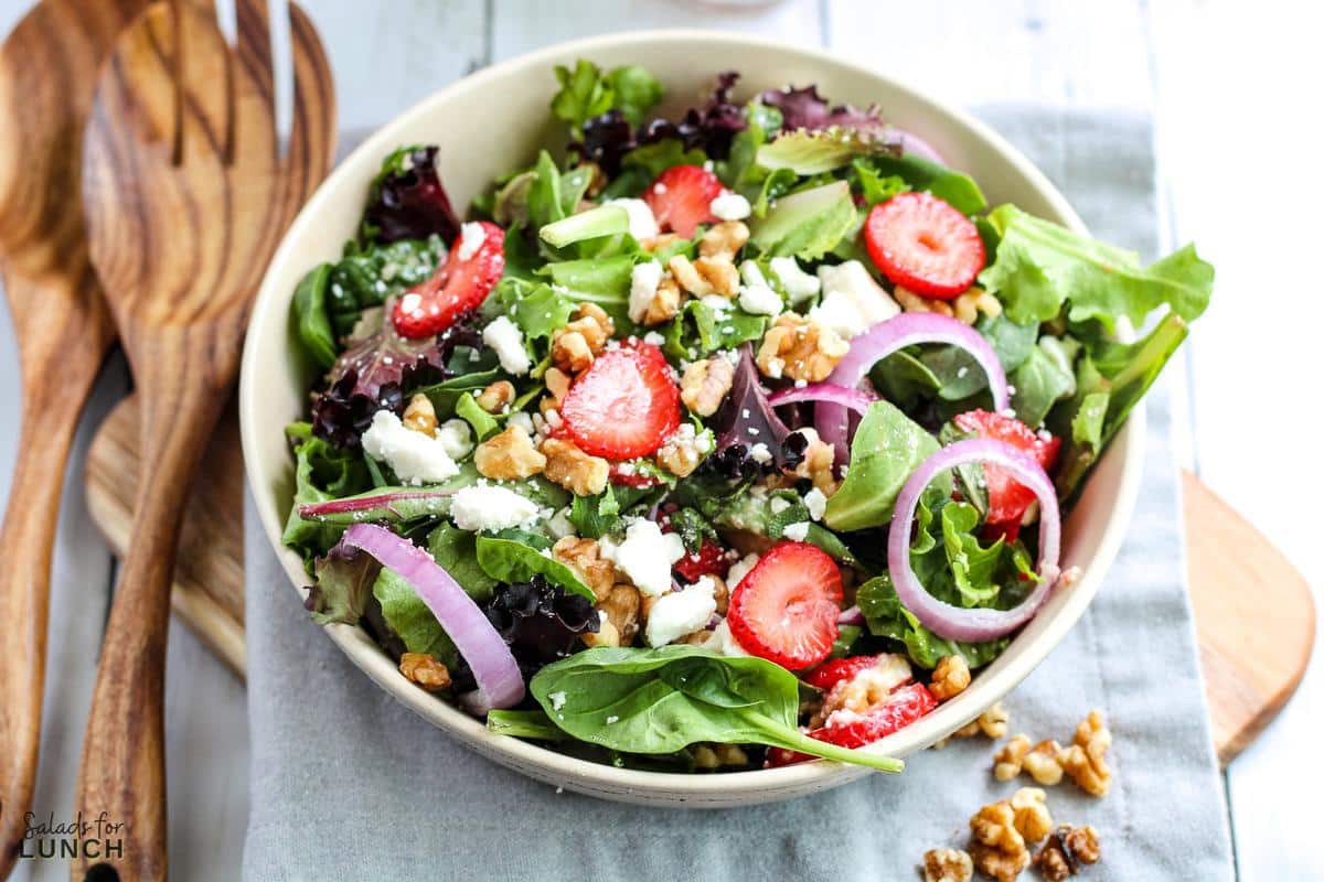 Overhead photo of a white bowl with a colourful strawberry, feta and mint salad with spring mix on a grey napkin.