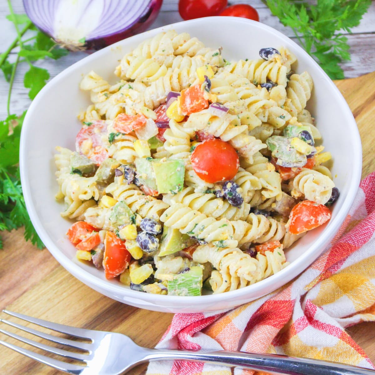 Square close up of a mexican pasta salad in a white bowl with black beans, cilantro, avocado, tomato, and corn.